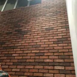 Ściana wyłożona dekoracyjną czerwoną cegłą z ciemnymi fugami, widok z dołu, fragment sufitu i okna na górze, worki z materiałami budowlanymi w lewym dolnym rogu.