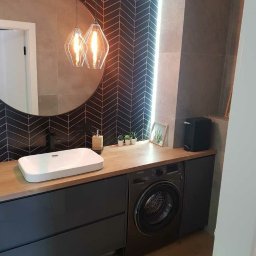 Usługi Remontowo - Budowlane Paweł Drywa - Nowoczesna łazienka z umywalką nablatową, pralką w zabudowie, lustrem w okrągłej ramie i dekoracyjną ścianą w ciemnoszarej jodełce ceramicznej.