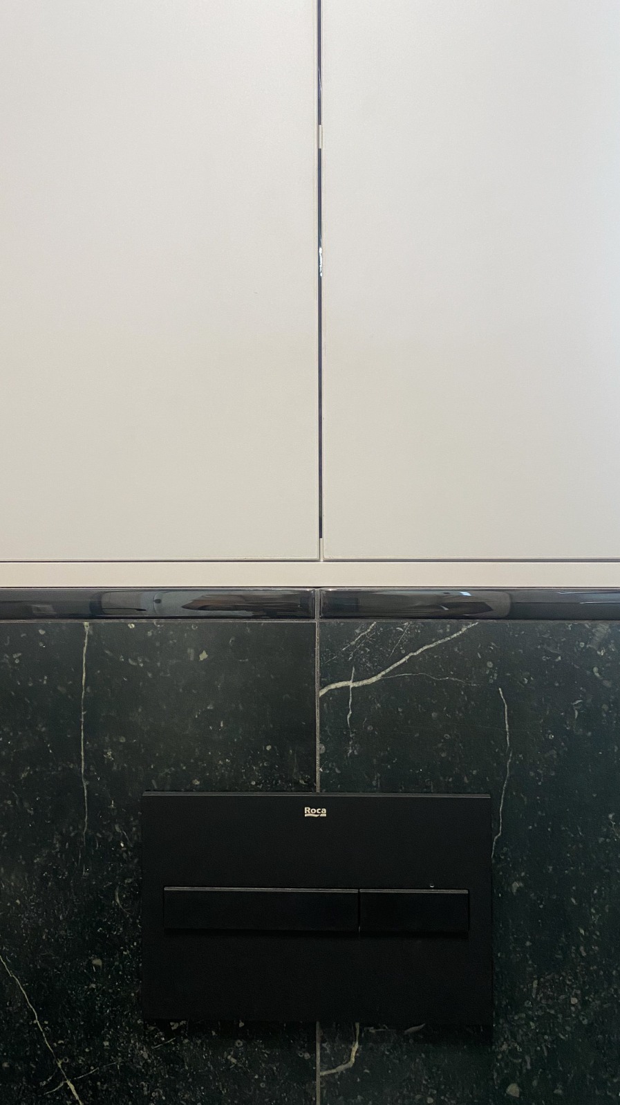 Fragment łazienki z czarnym marmurem z białymi żyłkami, białymi płytkami na górze i czarnym panelem spłukującym Roca.