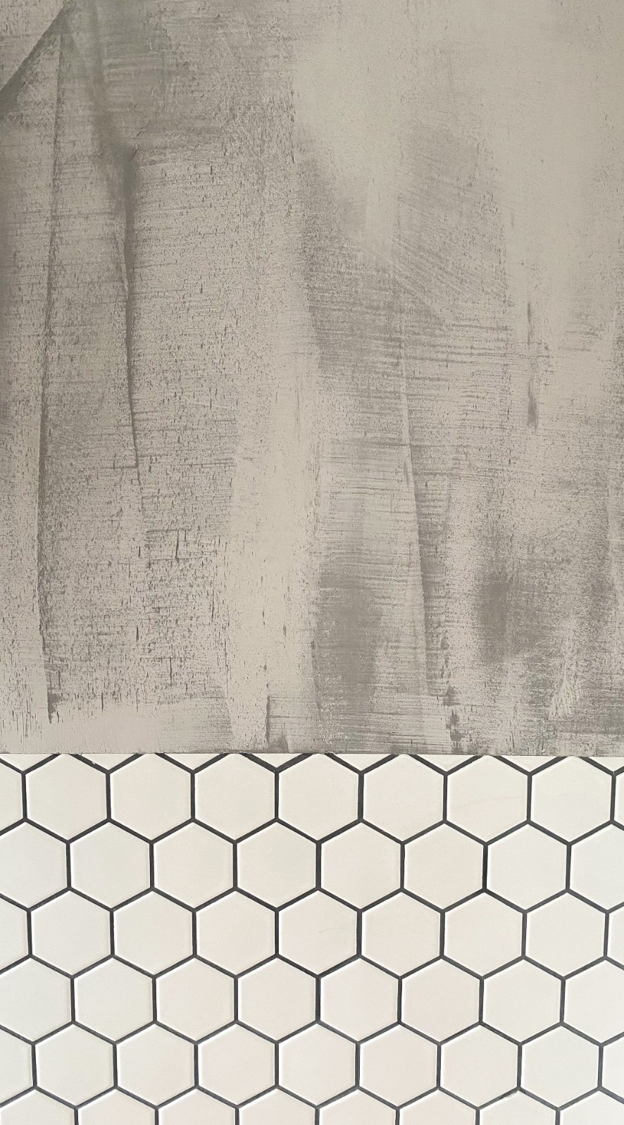 Fragment ściany z białymi heksagonalnymi płytkami i surowym, szarym wykończeniem powyżej, imitującym beton.