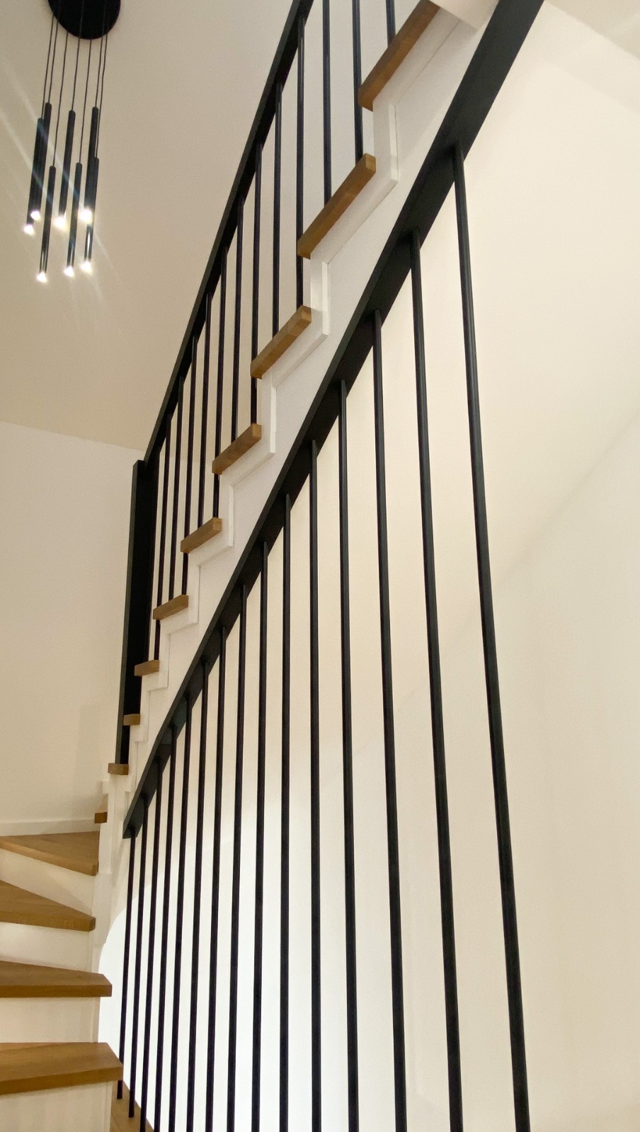 Nowoczesne schody z drewnianymi stopniami i czarną metalową balustradą, oświetlone przez designerską lampę wiszącą.