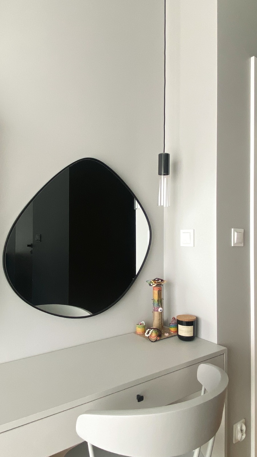 Nowoczesna łazienka z asymetrycznym lustrem w czarnej ramie, wiszącą lampą z przezroczystym kloszem i szarą toaletką z dekoracjami.