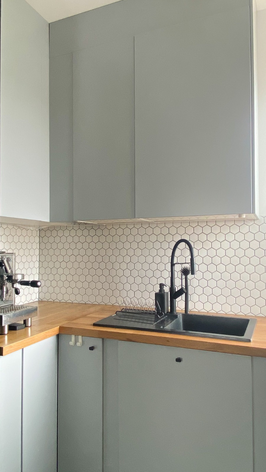 Szare szafki kuchenne z drewnianym blatem, heksagonalne płytki na ścianie, czarny zlew i bateria, ekspres do kawy na blacie po lewej stronie.