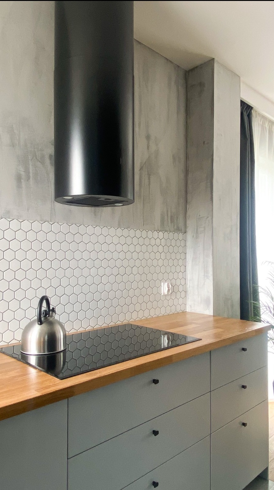 Nowoczesna kuchnia z szarymi szafkami, drewnianym blatem i heksagonalnymi płytkami na ścianie, uzupełniona czarnym okapem cylindrycznym i czajnikiem na płycie indukcyjnej.