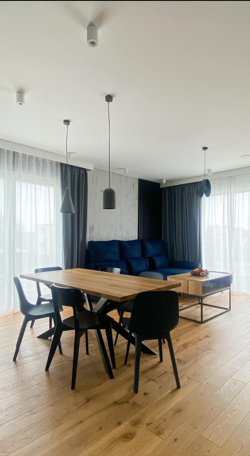 Jasne drewniane podłogi, stół z drewnianym blatem i czarnymi krzesłami, niebieska sofa w tle, szare zasłony i nowoczesne lampy wiszące.