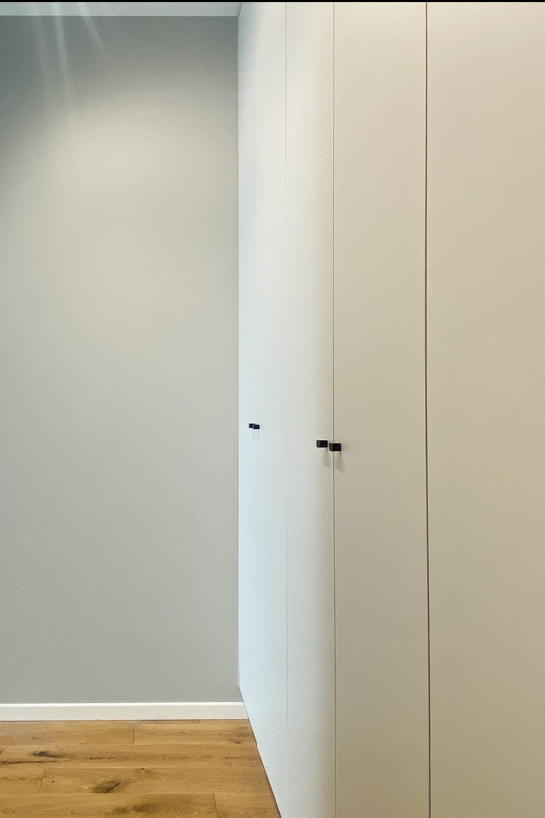 Minimalistyczna łazienka: Jasnoszara ściana kontrastuje z drewnianą podłogą i zabudowaną szafką z ciemnymi, kwadratowymi uchwytami.