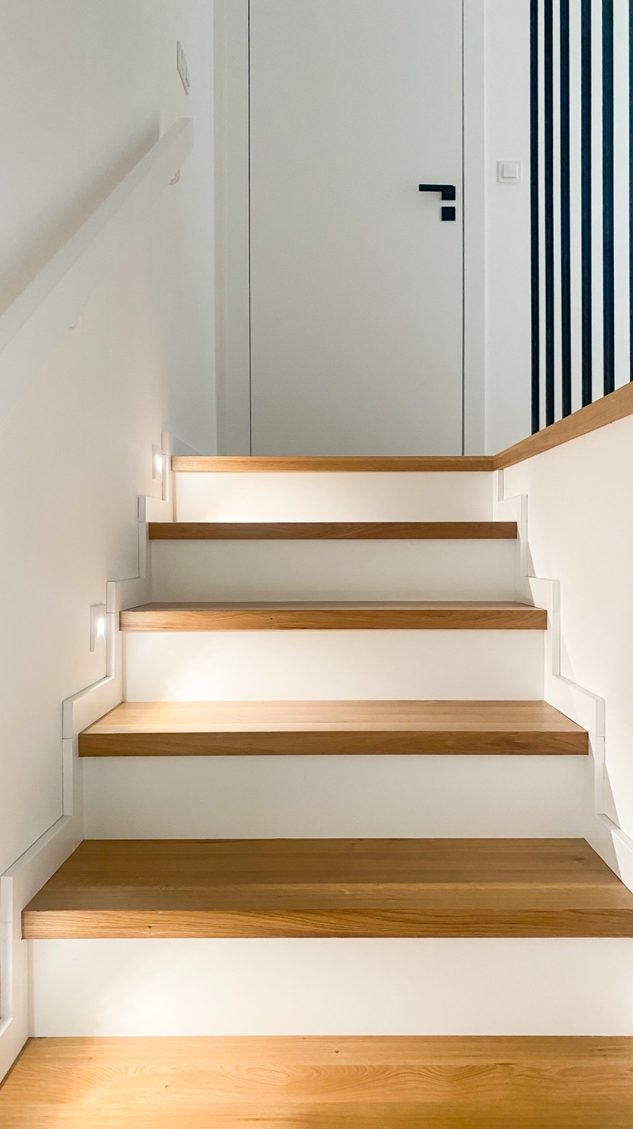 Nowoczesne schody drewniane z białymi podstopnicami i oświetleniem LED w ścianie, prowadzące do białych drzwi z czarną klamką i fragmentu ściany w czarno-białe pasy.