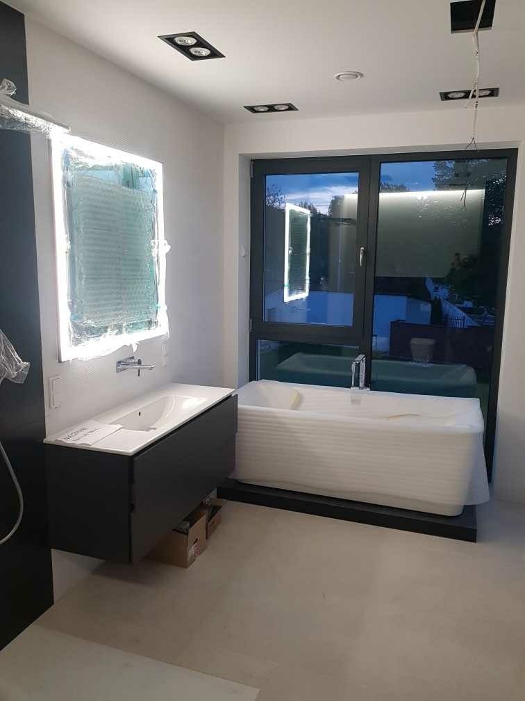 Nowoczesna łazienka w trakcie wykańczania: biała wanna obłożona folią ochronną stoi przy dużym oknie, obok szafka pod umywalkę w kolorze antracytowym, lustro z podświetleniem w folii, sufit...