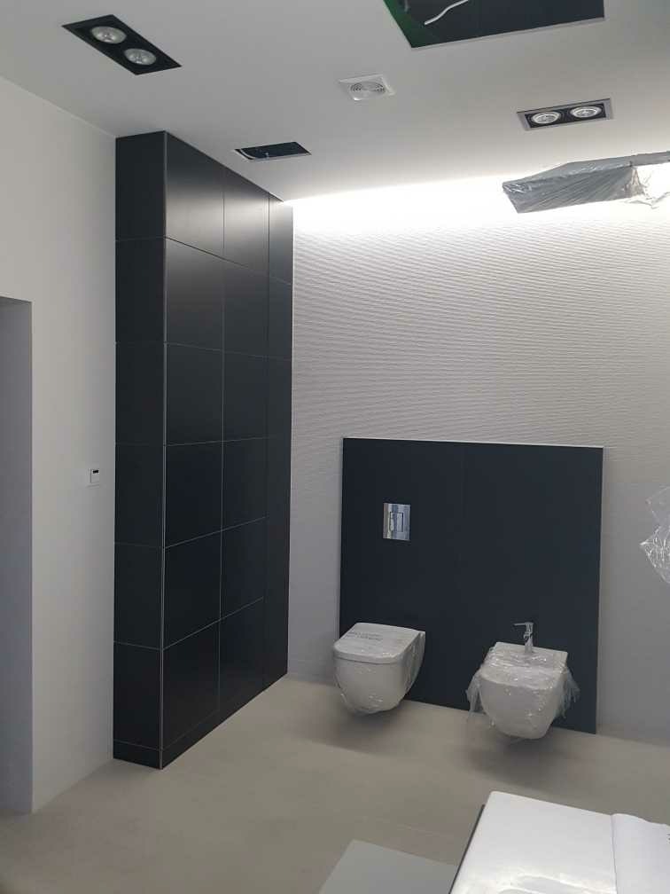 Nowoczesna łazienka w trakcie wykańczania: biała toaleta i bidet częściowo zabezpieczone folią, na tle czarnej ściany i białej ściany z delikatną fakturą, oświetlenie sufitowe.