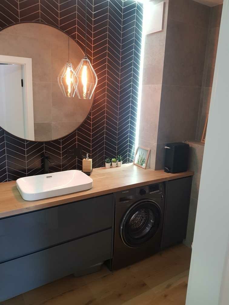 Nowoczesna łazienka z umywalką nablatową, pralką w zabudowie, lustrem w okrągłej ramie i dekoracyjną ścianą w ciemnoszarej jodełce ceramicznej.