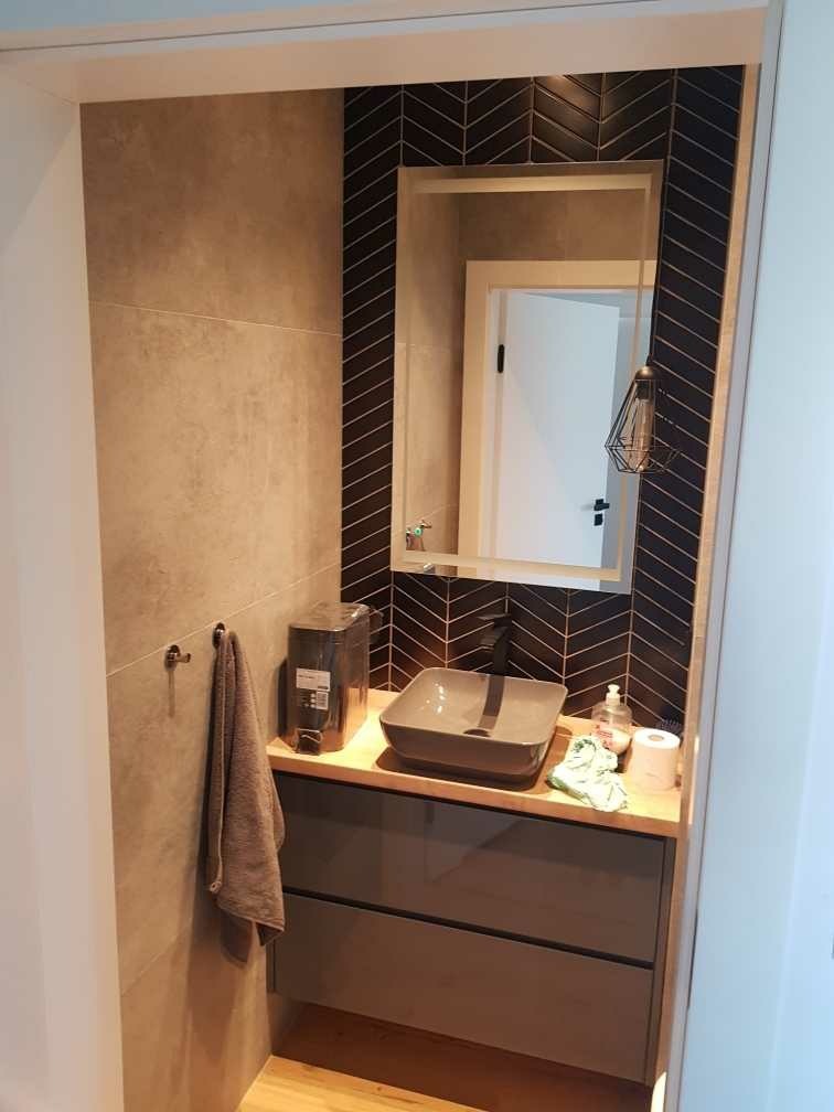 Nowoczesna łazienka z umywalką nablatową, czarną armaturą i płytkami ułożonymi w jodełkę za lustrem. Na blacie dozownik mydła i ręcznik papierowy. Na ścianie wisi ręcznik.