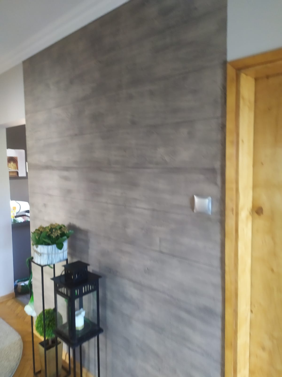 Ściana wykończona panelami imitującymi betonowe deski, z widocznym gniazdkiem elektrycznym i dekoracjami w postaci lampionu i kwiatów w tle, obok drewnianej ościeżnicy drzwi.