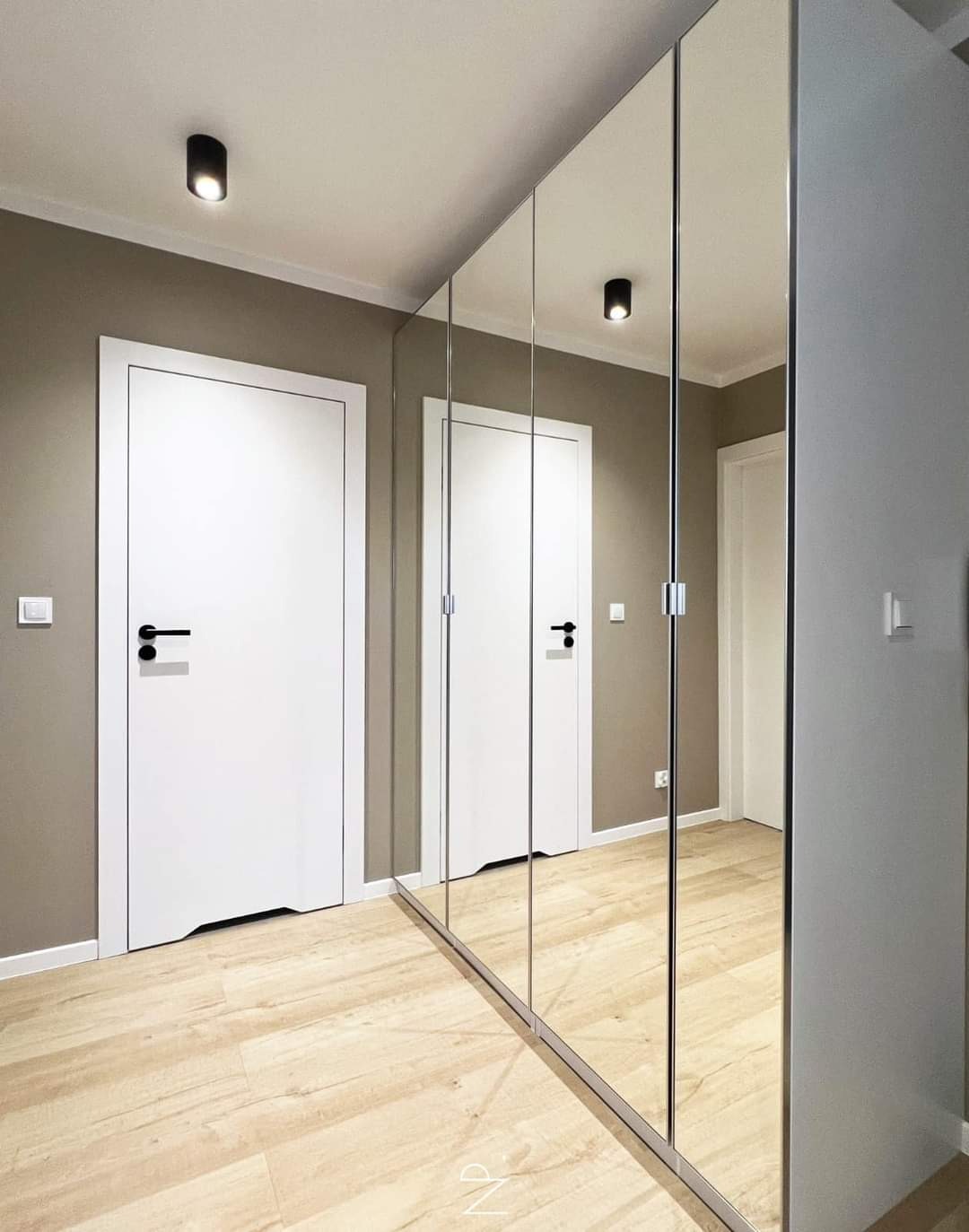Nowoczesne wnętrze: Lustrzana szafa przesuwna z białymi drzwiami i drewnianą podłogą w neutralnej kolorystyce, oświetlona punktowymi lampami sufitowymi.