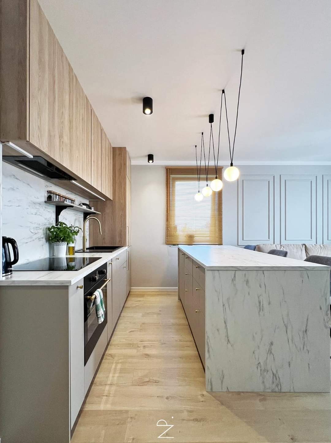Nowoczesna kuchnia w odcieniach szarości i drewna, z wyspą wykończoną marmurem, oświetlona wiszącymi lampami kulami i punktowymi reflektorami, z widocznym fragmentem salonu w tle.