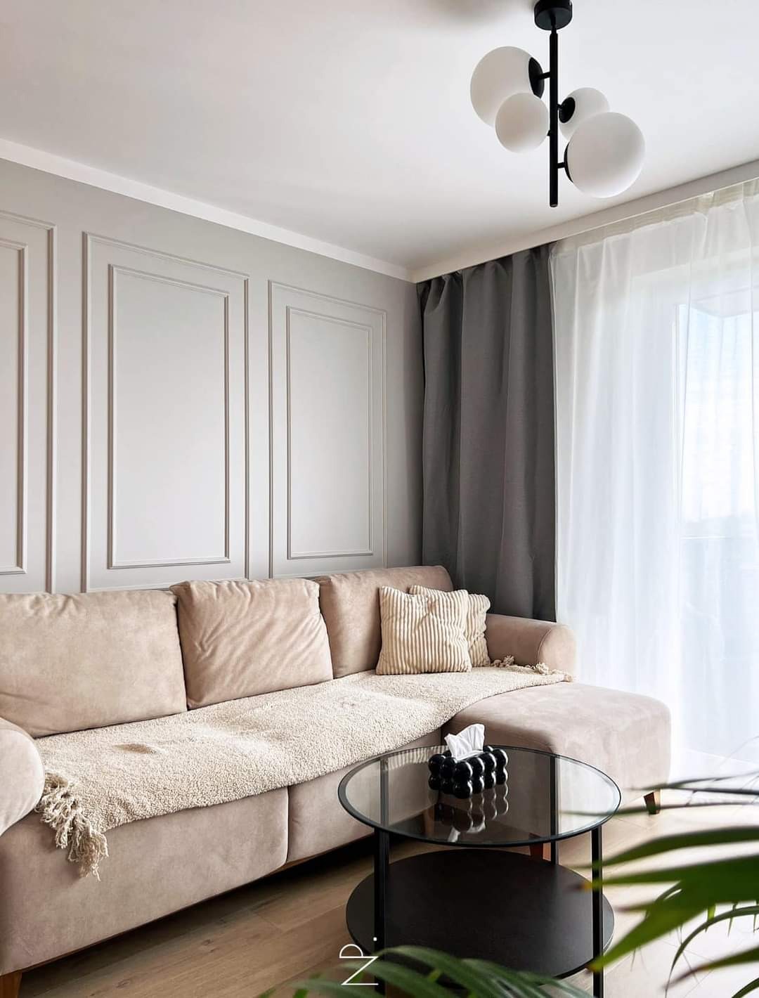 Jasny salon z beżową sofą, okrągłym szklanym stolikiem kawowym i panelami ściennymi w stylu klasycystycznym, oświetlony nowoczesną lampą sufitową.