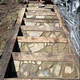 Bąbiak Kamil Usługi Budowlane - Budowa schodów z kamienia naturalnego w drewnianej ramie, widoczny pracownik z narzędziami i wiadro z zaprawą.