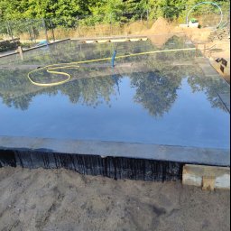 Bąbiak Kamil Usługi Budowlane - Przygotowany teren pod wylewkę betonową, widoczna folia hydroizolacyjna, żółty wąż ogrodowy i odbicie drzew w wodzie na powierzchni betonu.