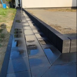 Bąbiak Kamil Usługi Budowlane - Świeżo ułożona, mokra, czarna kostka brukowa przy białym budynku z szarymi oknami, widoczne ślady po deszczu.