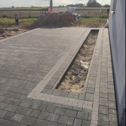 Bąbiak Kamil Usługi Budowlane - Częściowo ułożona, szara kostka brukowa z jasnymi pasami dekoracyjnymi; widoczny rów z piaskiem i kablami, oraz kopiec piasku w tle z dwiema osobami.