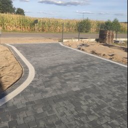 Bąbiak Kamil Usługi Budowlane - Nowa, ciemnoszara nawierzchnia z kostki brukowej z jasnoszarą obwódką, otoczona piaskiem i z widocznym polem kukurydzy w tle.