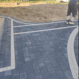 Bąbiak Kamil Usługi Budowlane - Częściowo ułożona, ciemnoszara kostka brukowa z jasnymi, wąskimi liniami oddzielającymi, widoczny fragment przygotowanego podłoża z piasku, w tle samochód dostawczy.