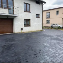 Ułożona ciemnoszara kostka brukowa na podjeździe przed domem z brązową bramą garażową i balkonem ze srebrną balustradą, widoczne ślady wilgoci na kostce.