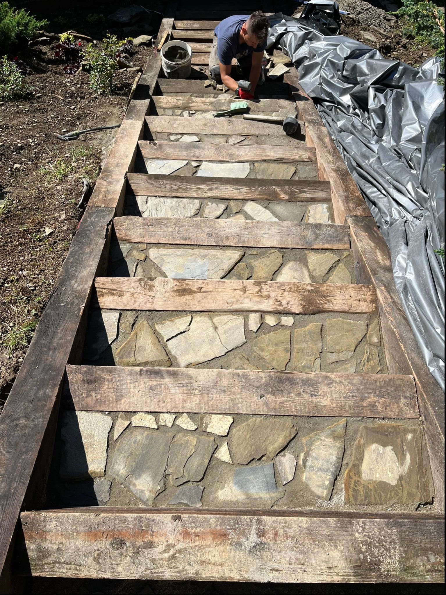 Budowa schodów z kamienia naturalnego w drewnianej ramie, widoczny pracownik z narzędziami i wiadro z zaprawą.