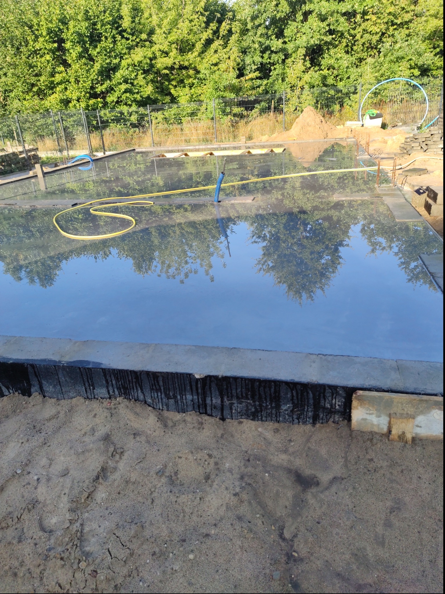 Przygotowany teren pod wylewkę betonową, widoczna folia hydroizolacyjna, żółty wąż ogrodowy i odbicie drzew w wodzie na powierzchni betonu.