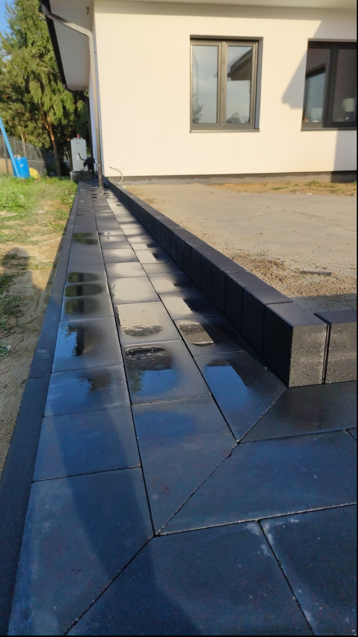 Świeżo ułożona, mokra, czarna kostka brukowa przy białym budynku z szarymi oknami, widoczne ślady po deszczu.