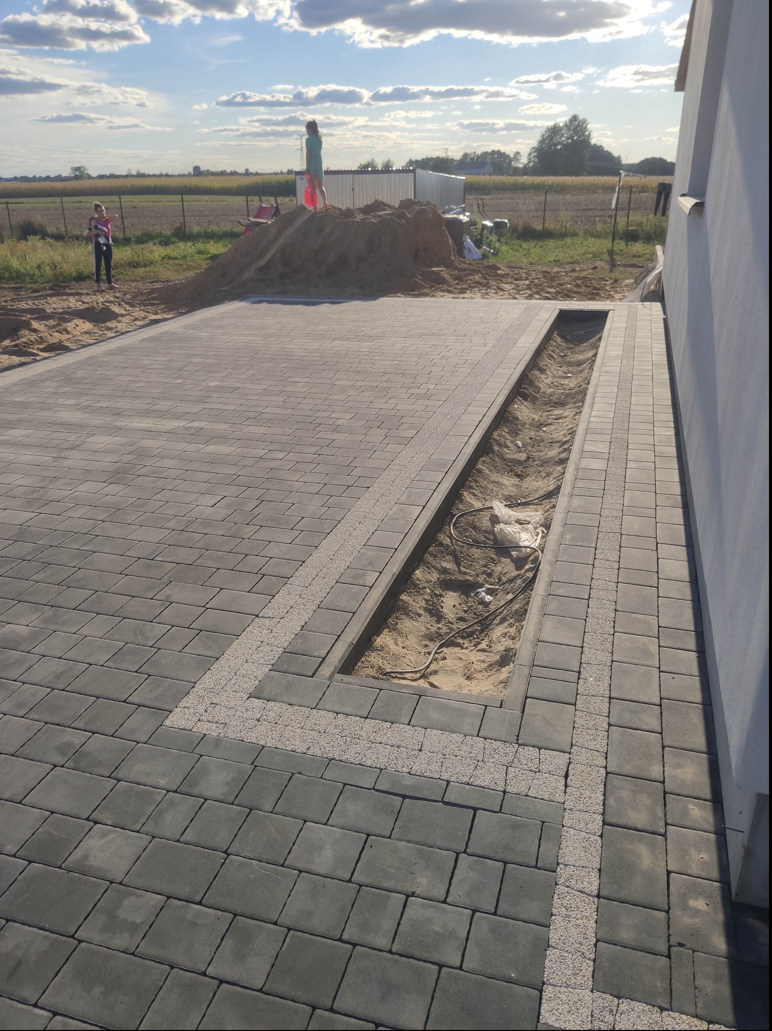 Częściowo ułożona, szara kostka brukowa z jasnymi pasami dekoracyjnymi; widoczny rów z piaskiem i kablami, oraz kopiec piasku w tle z dwiema osobami.