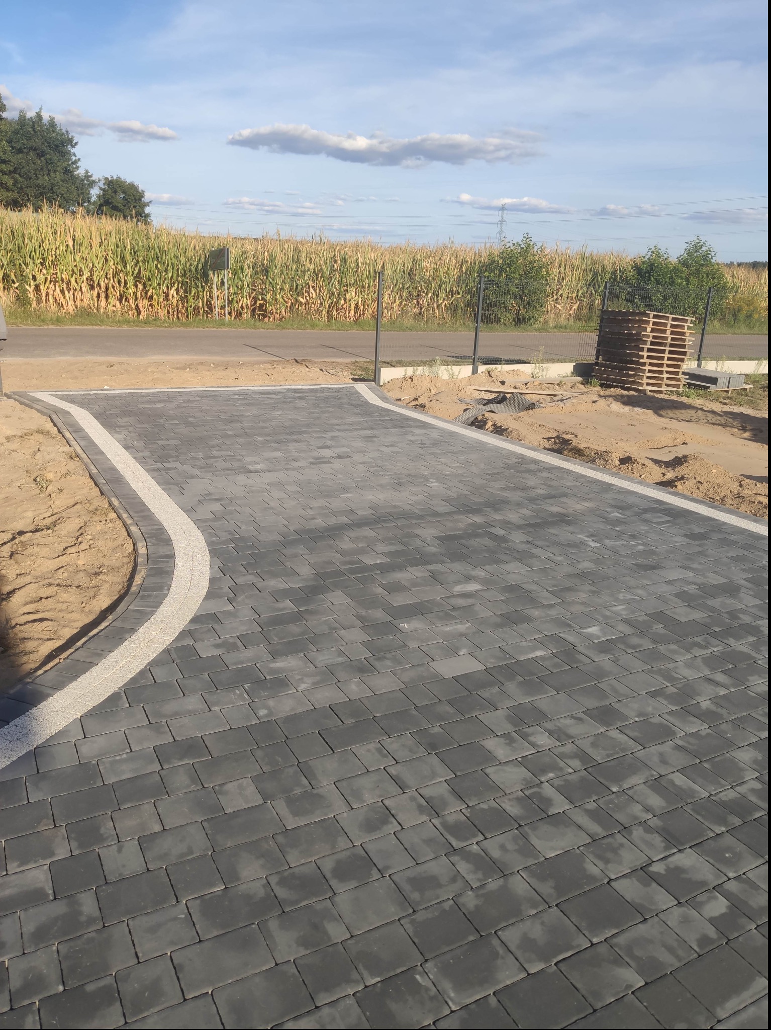 Nowa, ciemnoszara nawierzchnia z kostki brukowej z jasnoszarą obwódką, otoczona piaskiem i z widocznym polem kukurydzy w tle.