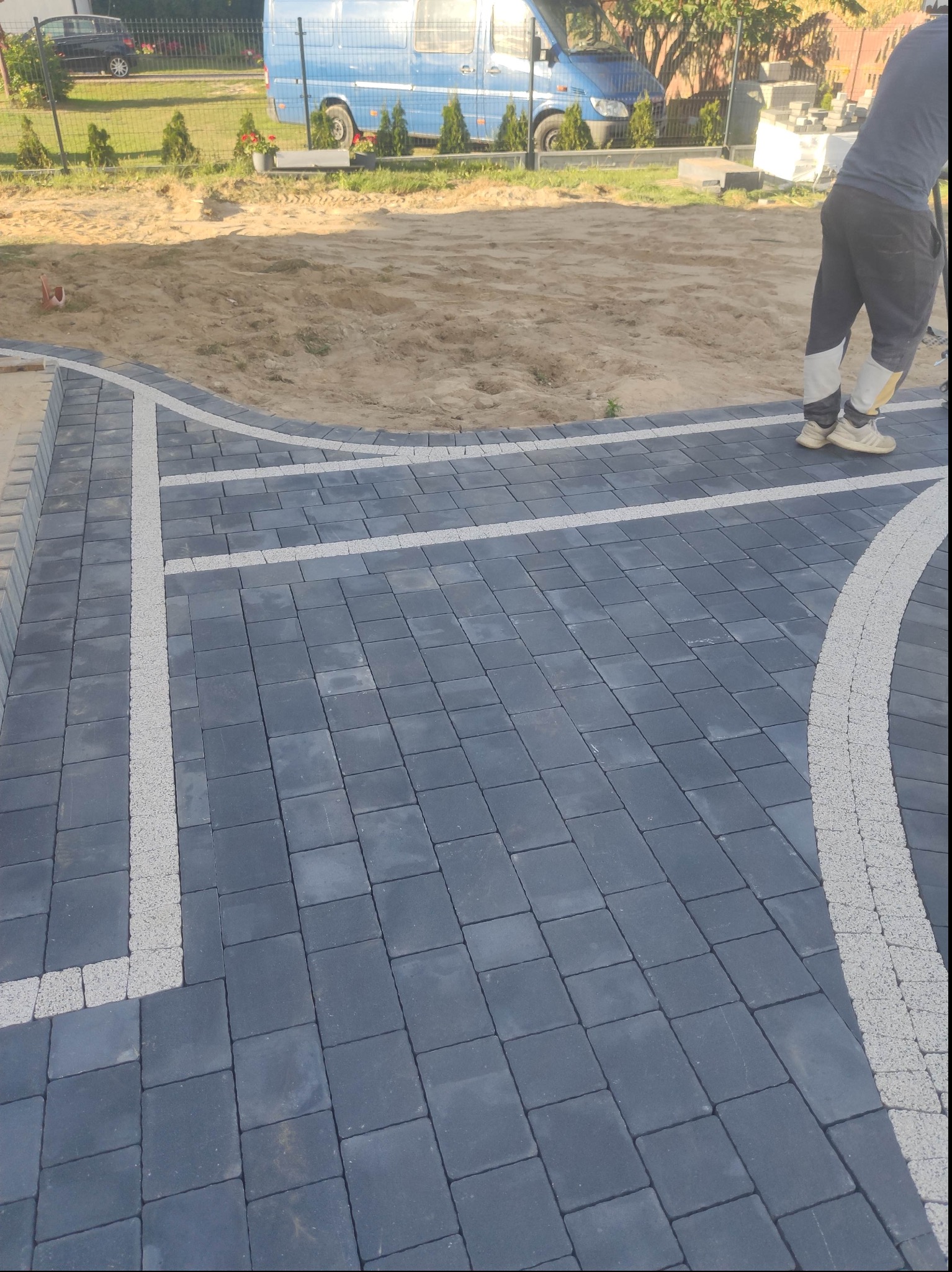 Częściowo ułożona, ciemnoszara kostka brukowa z jasnymi, wąskimi liniami oddzielającymi, widoczny fragment przygotowanego podłoża z piasku, w tle samochód dostawczy.