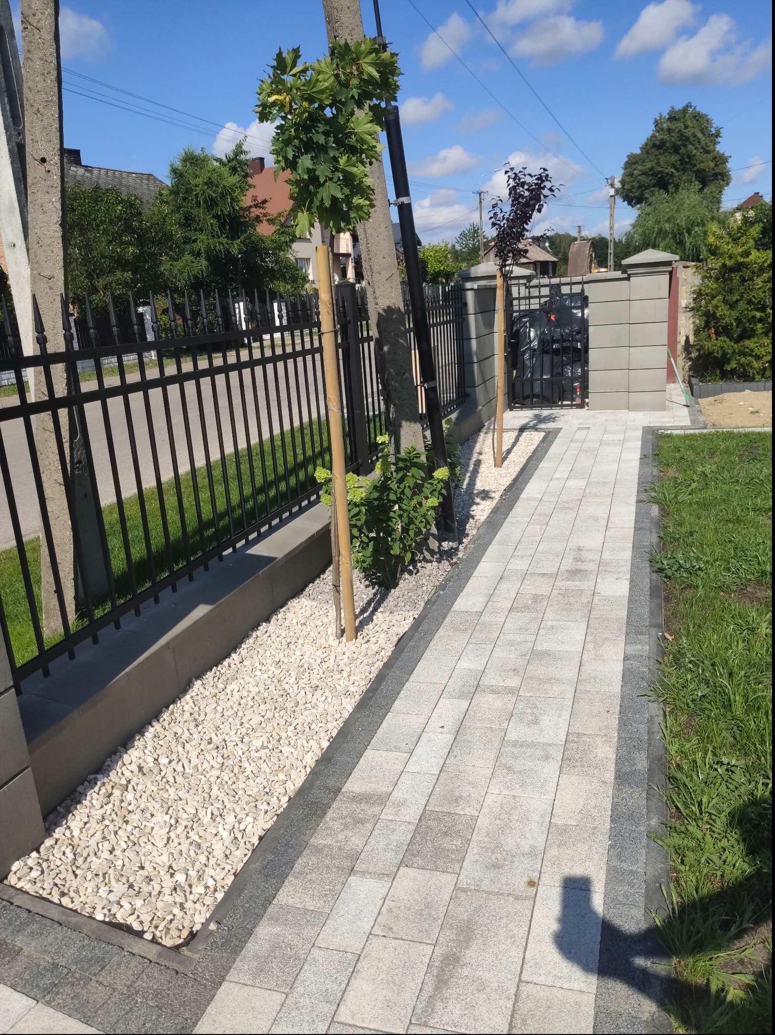 Nowoczesna ścieżka z jasnoszarej kostki brukowej obramowana ciemniejszą kostką, z czarnym metalowym ogrodzeniem i rabatą z białego kamienia, małe drzewka posadzone wzdłuż ogrodzenia.