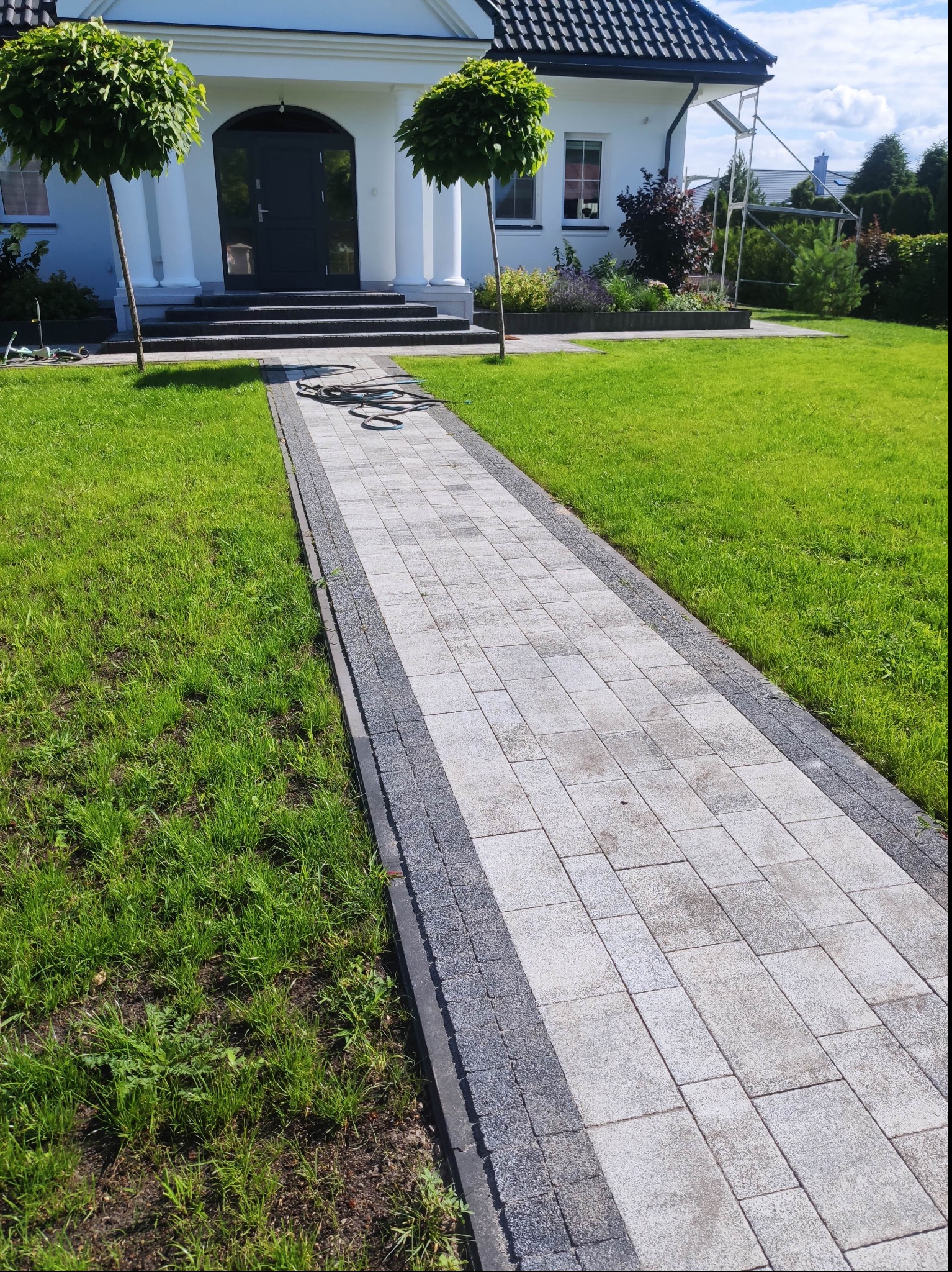Chodnik z jasnoszarej kostki brukowej z ciemnoszarym obrzeżem prowadzący do wejścia do białego domu z kolumnami, na trawniku zwinięty wąż ogrodowy.