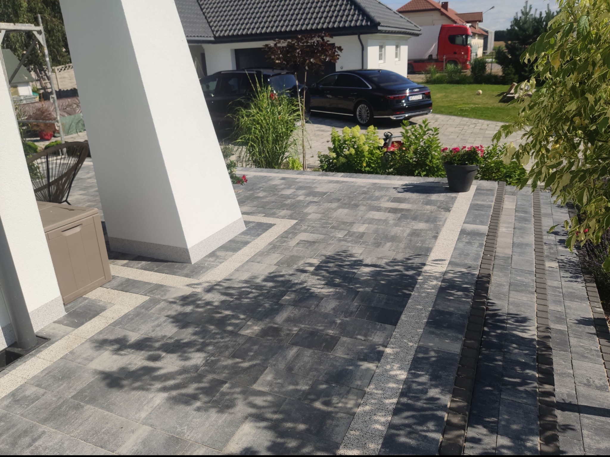 Nowoczesny podjazd z szarej kostki brukowej ułożonej w geometryczny wzór, z jasnymi paskami dekoracyjnymi, prowadzący do domu z dwoma czarnymi samochodami w tle, cień drzewa na nawierzchni.