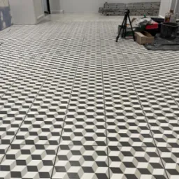 Ułożona podłoga z płytek w geometryczny wzór 3D w odcieniach szarości i bieli, widoczne akcesoria glazurnicze, drabina oraz nie wykończona ściana w tle.