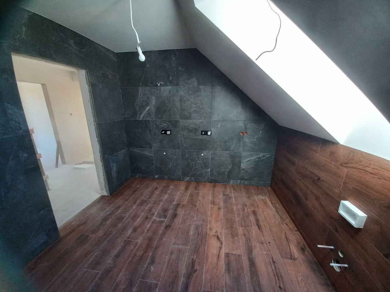 Łazienka na poddaszu w trakcie remontu: ciemnoszare płytki imitujące kamień na ścianach i drewnopodobne panele na podłodze, widoczne punkty elektryczne, surowa żarówka i fragment okna dachowego.