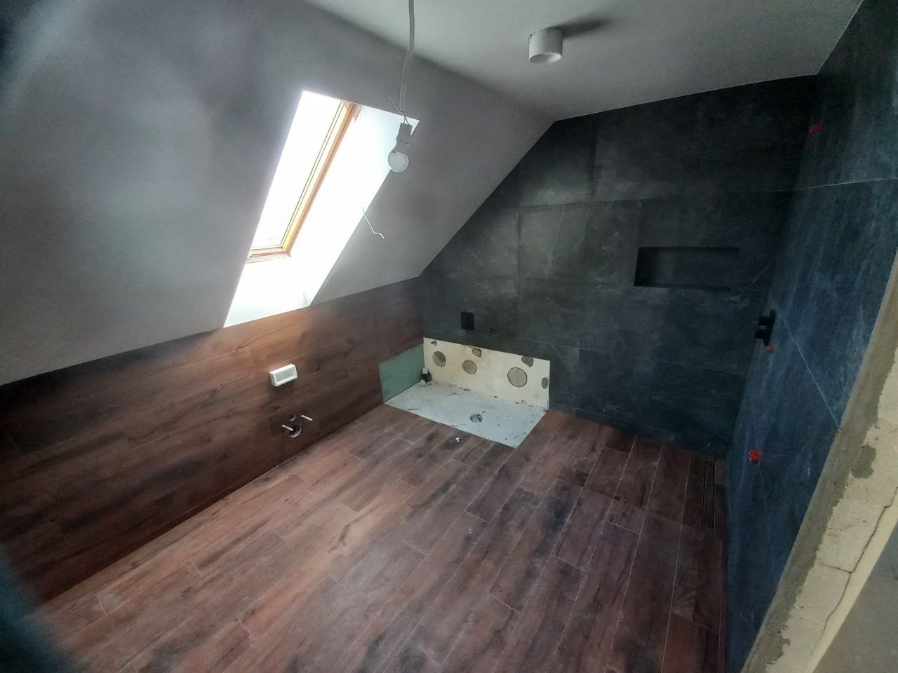 Łazienka w trakcie wykańczania na poddaszu: ciemne płytki imitujące kamień na ścianie z wnęką, drewnopodobne płytki na podłodze i ścianie pod oknem dachowym, widoczne instalacje hydrauliczne.