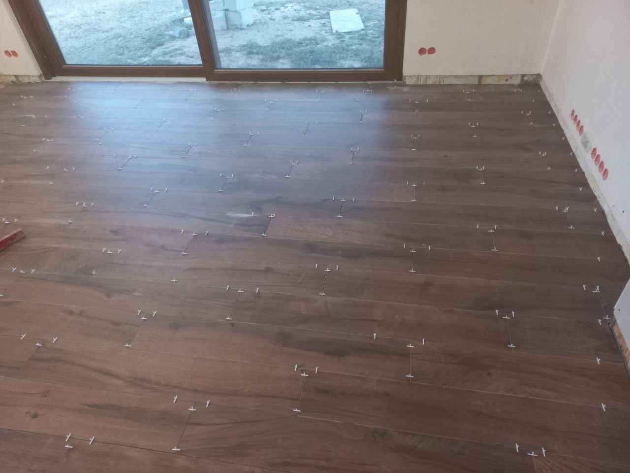 Układanie płytek imitujących drewno na podłodze, widoczne krzyżyki dystansowe, przygotowanie do fugowania przy oknie i ścianie.