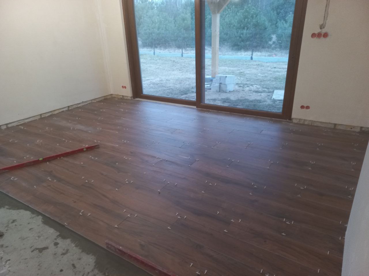 Układanie brązowych płytek imitujących drewno na podłodze, widoczne krzyżyki dystansowe, poziomica i surowy beton w rogu pomieszczenia przed dużym oknem.