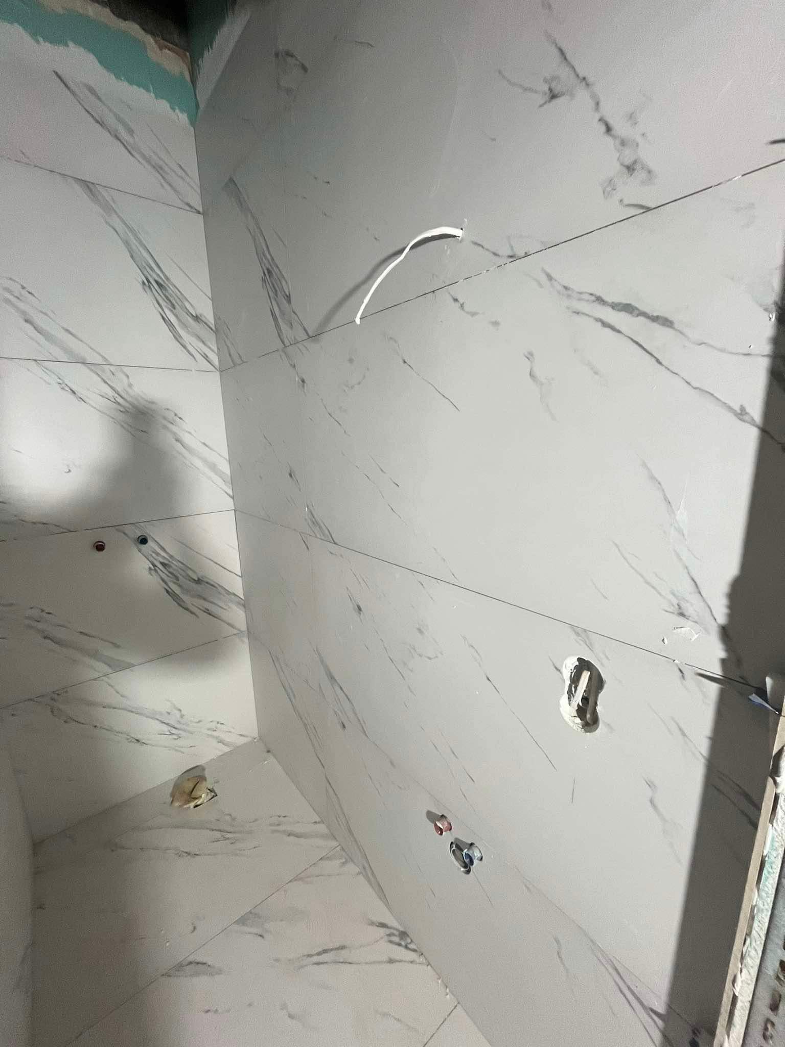 Ściany i podłoga w trakcie układania płytek imitujących marmur w łazience, widoczne otwory na rury i kable, przygotowane pod białą armaturę.