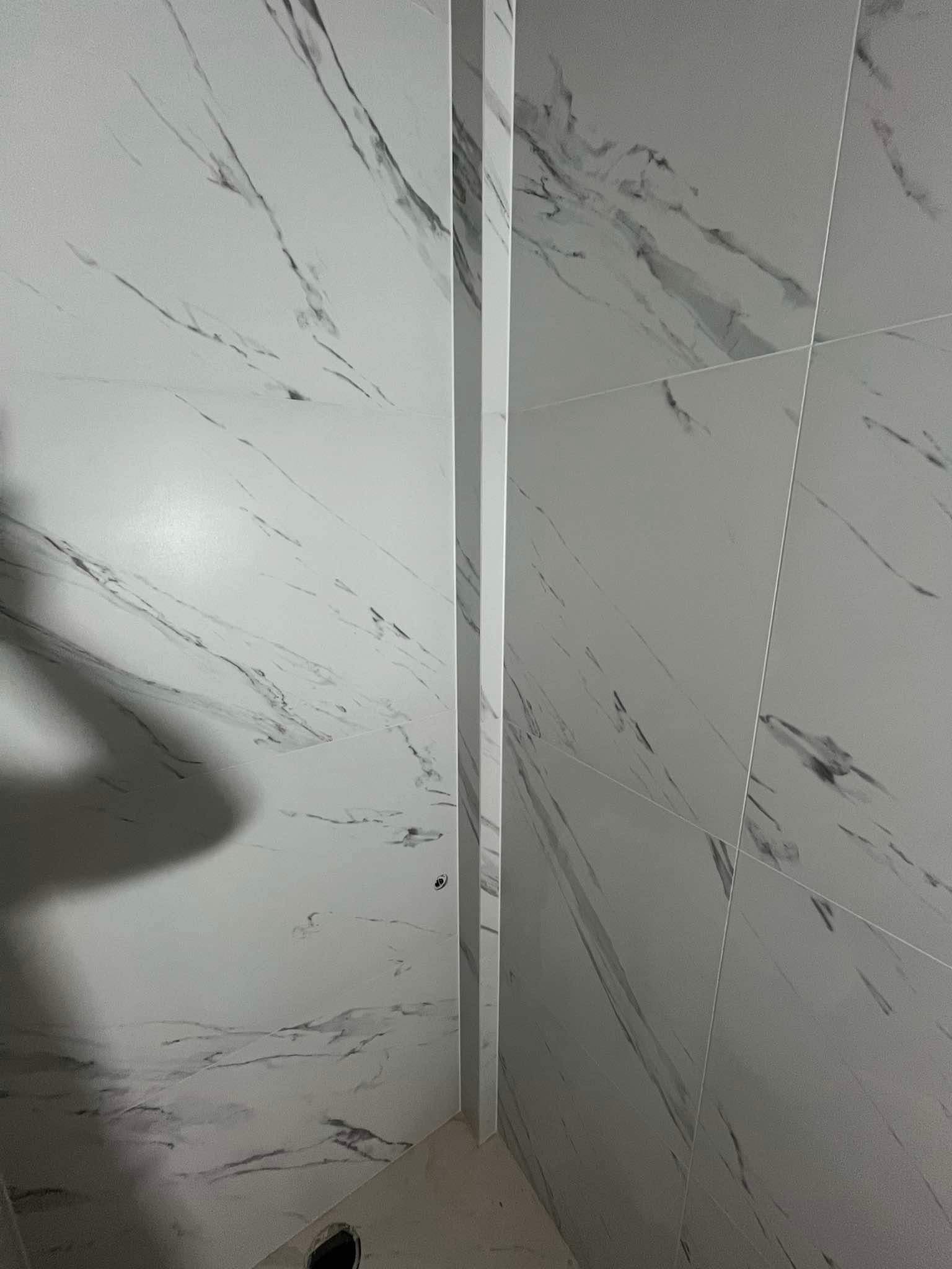 Ściana w łazience wyłożona białymi płytkami imitującymi marmur z wyraźnymi szarymi żyłami, widoczna listwa narożna i otwór w podłodze.