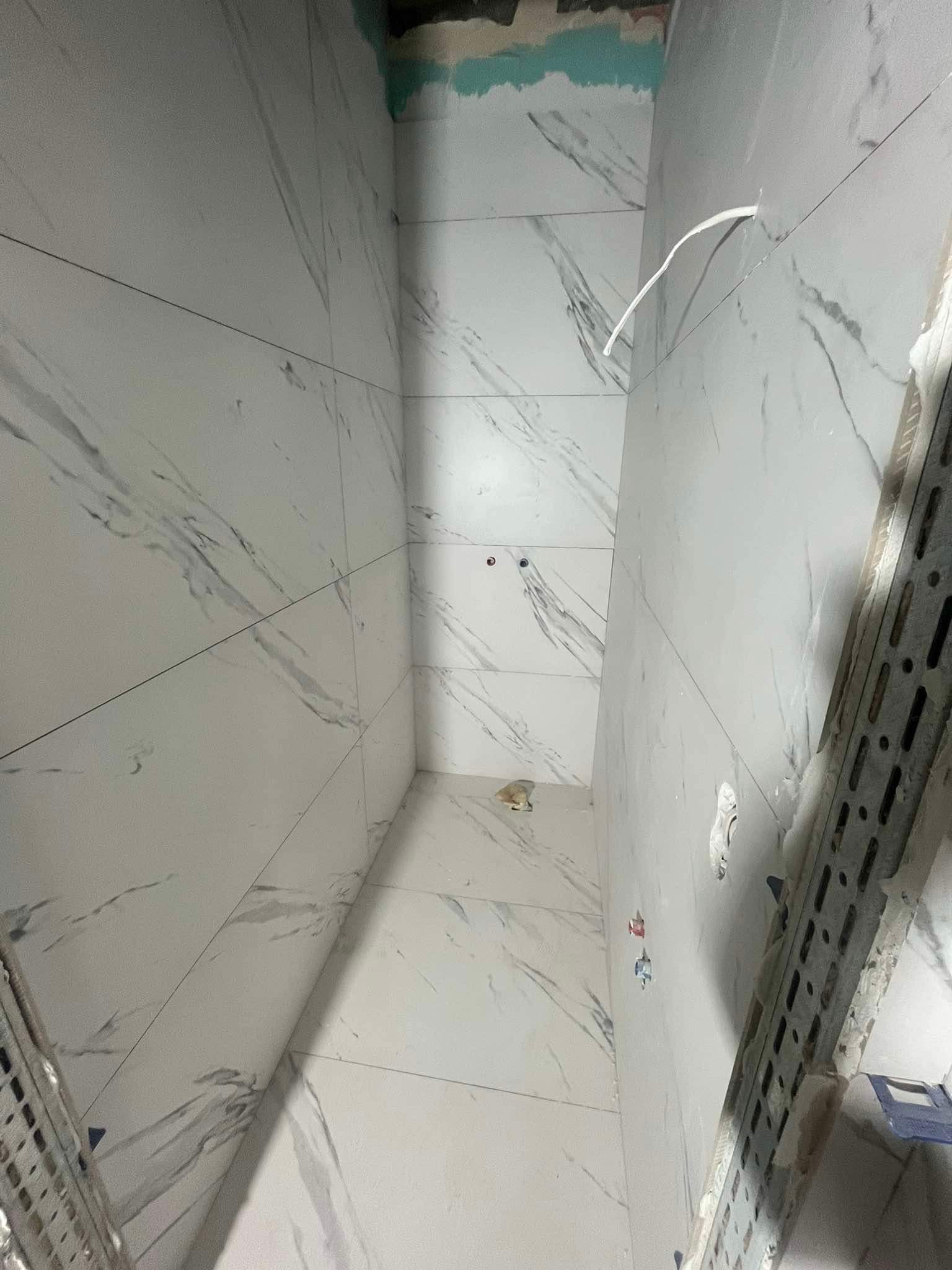 Świeżo ułożone, białe płytki imitujące marmur na ścianach i podłodze w małej łazience w trakcie remontu. Widoczne wystające kable i fragment rusztowania.