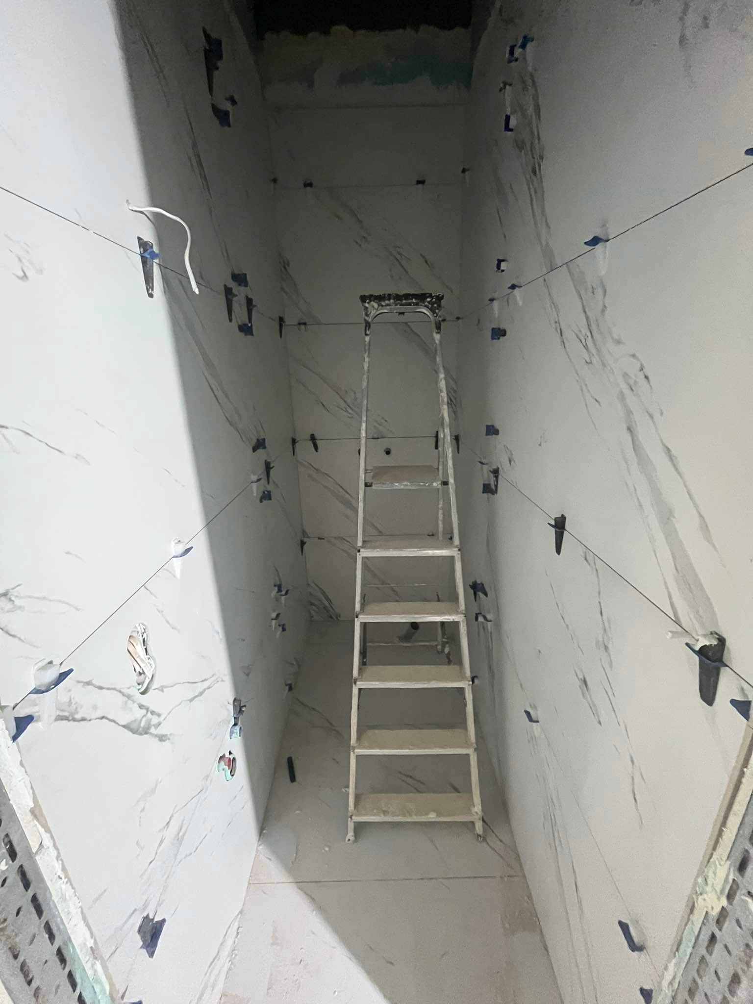 Wąskie wnętrze w trakcie układania marmurowych płytek ściennych, widoczna drabina, sznurki poziomujące z klipsami i krzyżyki dystansowe.