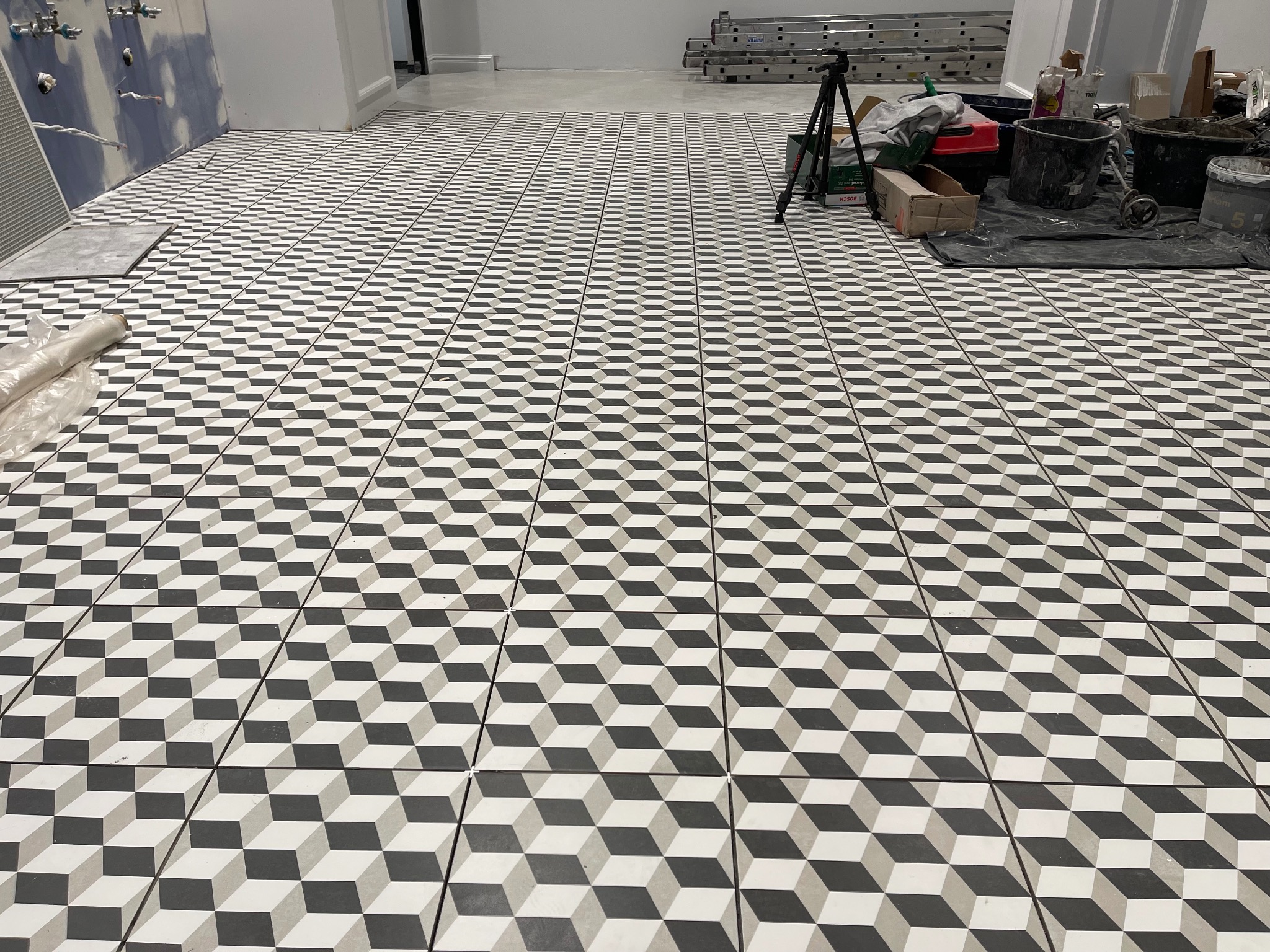 Ułożona podłoga z płytek w geometryczny wzór 3D w odcieniach szarości i bieli, widoczne akcesoria glazurnicze, drabina oraz nie wykończona ściana w tle.
