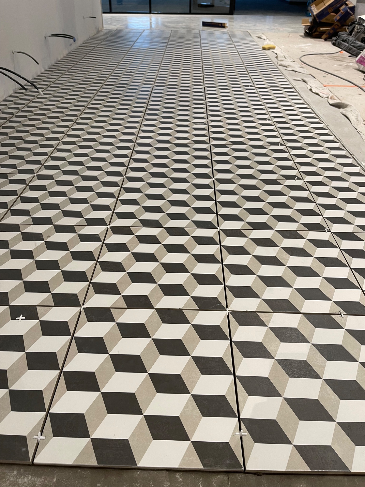 Ułożona podłoga z płytek o wzorze 3D w odcieniach szarości i bieli, widoczne krzyżyki dystansowe, surowy stan wykończenia.