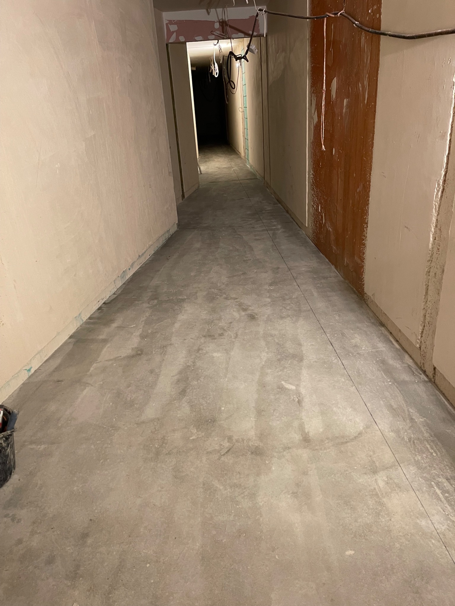Długi korytarz w trakcie remontu: surowy beton na podłodze, otynkowane ściany z widocznymi śladami prac, linia wyznaczająca poziom na podłodze, widoczne instalacje pod sufitem.