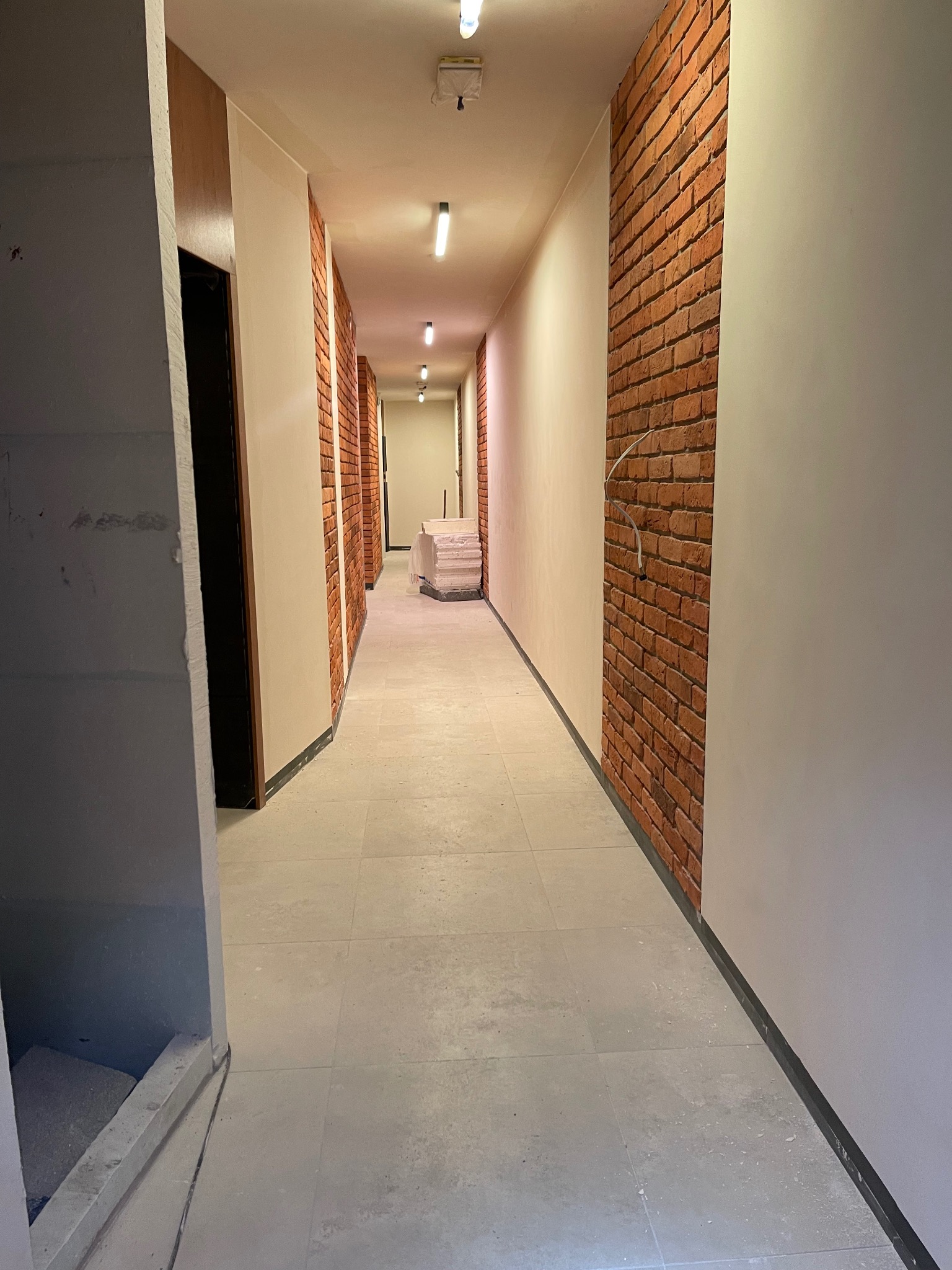 Korytarz w trakcie wykańczania, z fragmentem ściany wyłożonym czerwoną cegłą, szare płytki na podłodze i białe ściany. Widoczne przewody wystające ze ściany z cegły.