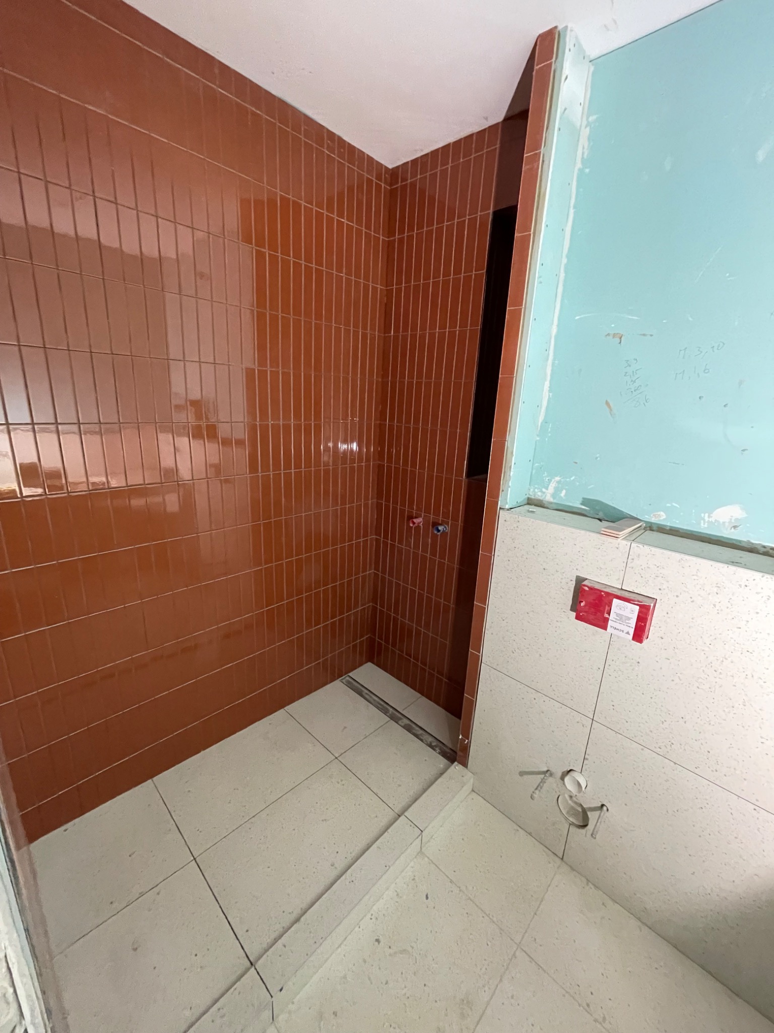 Wnętrze łazienki w trakcie wykańczania: brązowe, pionowo ułożone płytki na ścianie prysznica, białe płytki podłogowe i fragment ściany z niebieską farbą oraz instalacją podtynkową.