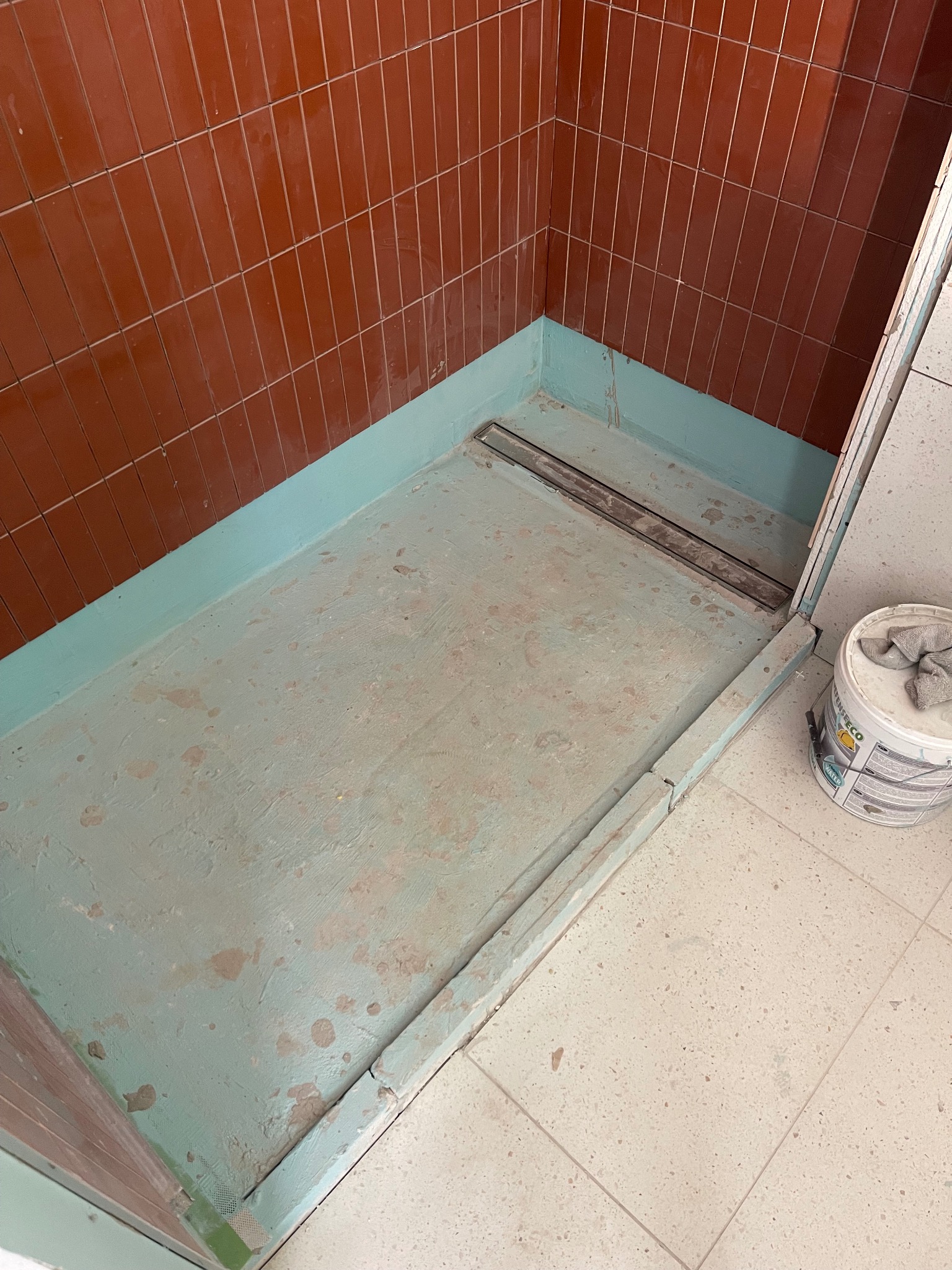 Niedokończony brodzik z odpływem liniowym, pomalowany na zielono, z brązowymi płytkami na ścianach, widoczny pojemnik z farbą i szmatką.